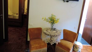 Ein Zimmer mit zwei Sesseln, einem kleinen Tisch mit Blumenvase, einem an der Wand montierten Fernseher und einem Holzschrank.