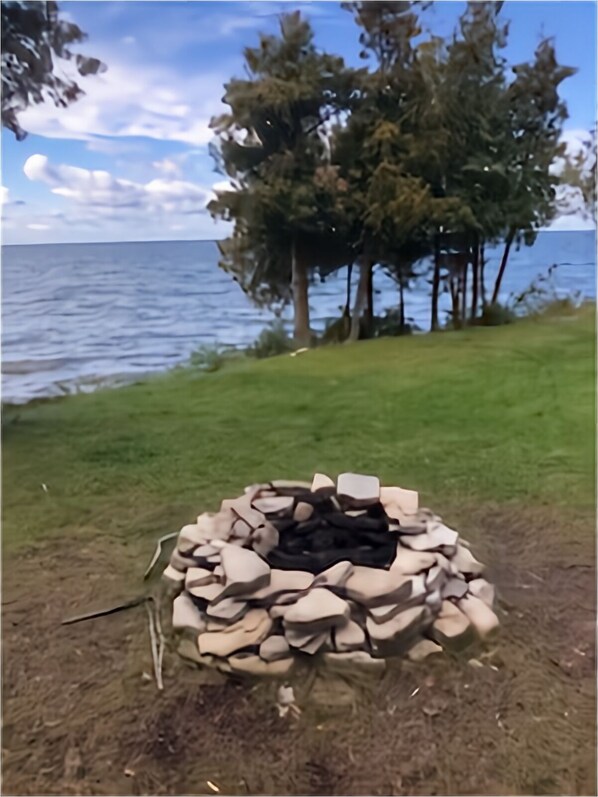 A campfire pit surrounded by stones, with a grassy area and trees in the background. - Waterfront Log Home, WiFi, 3 bedrooms, Direct TV, Fire Pit (Egg Harbor)