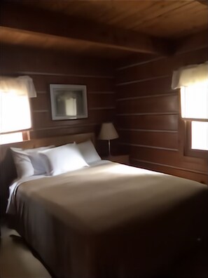 A wooden cabin room with a bed, a nightstand, a lamp, and a window with curtains. - Waterfront Log Home, WiFi, 3 bedrooms, Direct TV, Fire Pit (Egg Harbor)