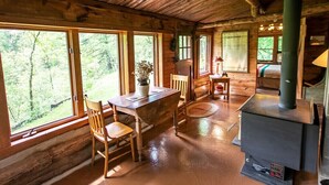 Ein rustikales Blockhaus-Interieur mit einem Holzespeisetisch, Stühlen, einem Herd und großen Fenstern mit Blick auf den Wald.