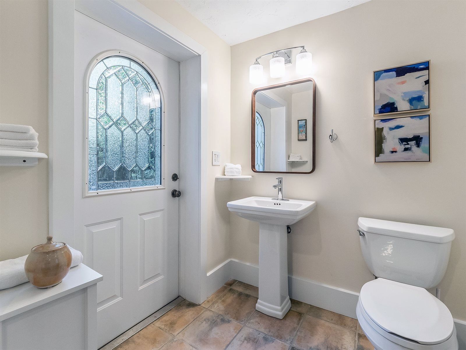 Un baño con una puerta blanca, un lavamanos blanco, un inodoro y una ventana con un diseño geométrico.