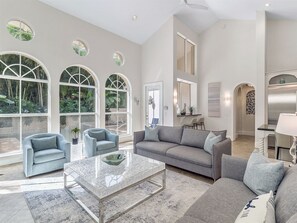 A spacious living room with a grey sofa, two armchairs, a marble coffee table, and large arched windows offering a view of greenery.