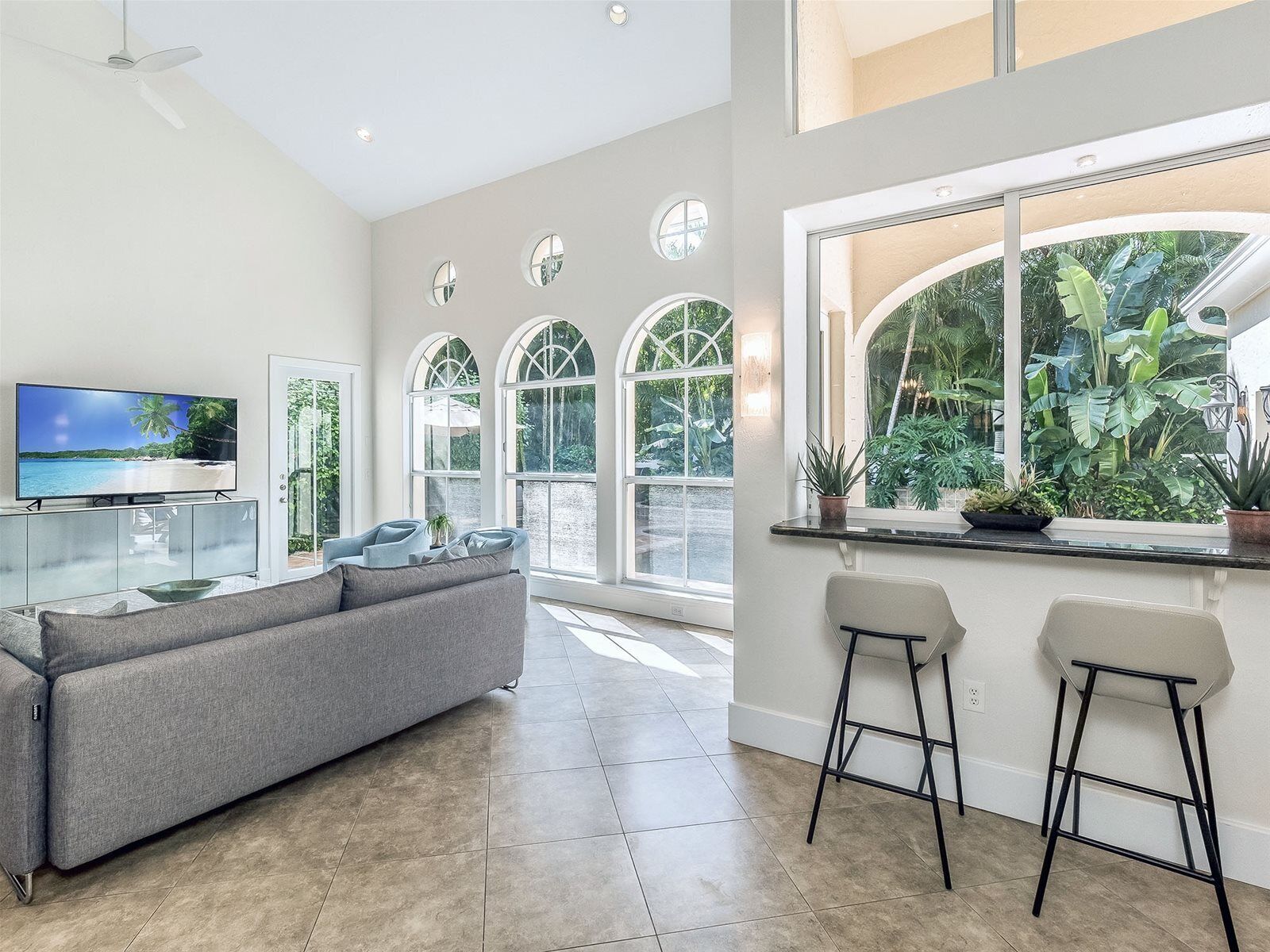 A spacious living room with a grey sofa, a flat-screen TV, and a large window overlooking a tropical garden.