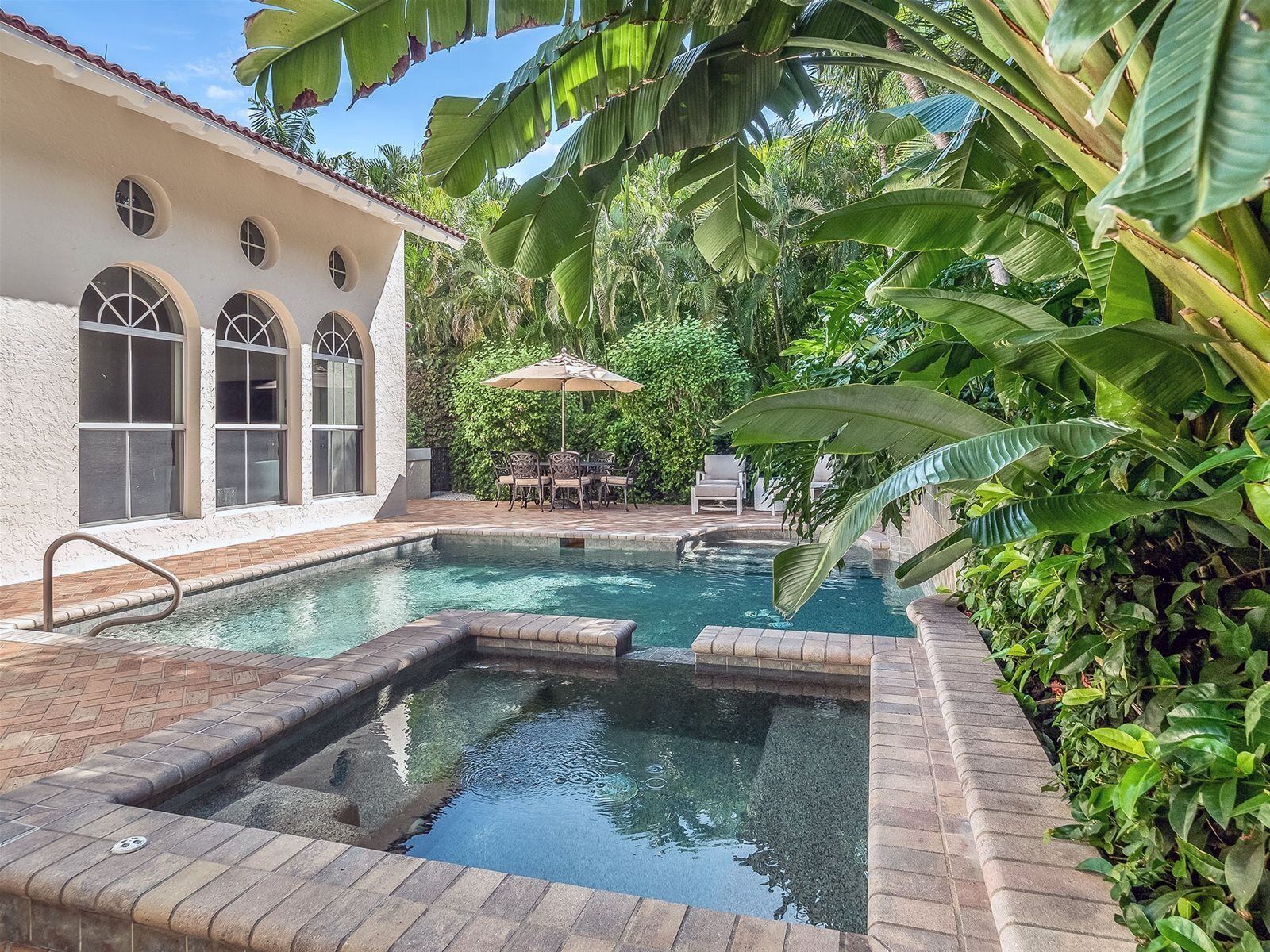 A swimming pool with a brick border, surrounded by lush greenery and a white building with arched windows.