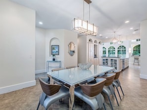 A spacious dining area with a large glass table surrounded by modern chairs, a pendant light, and a view of the kitchen and garden through large windows. - Casa Captiva (Captiva)
