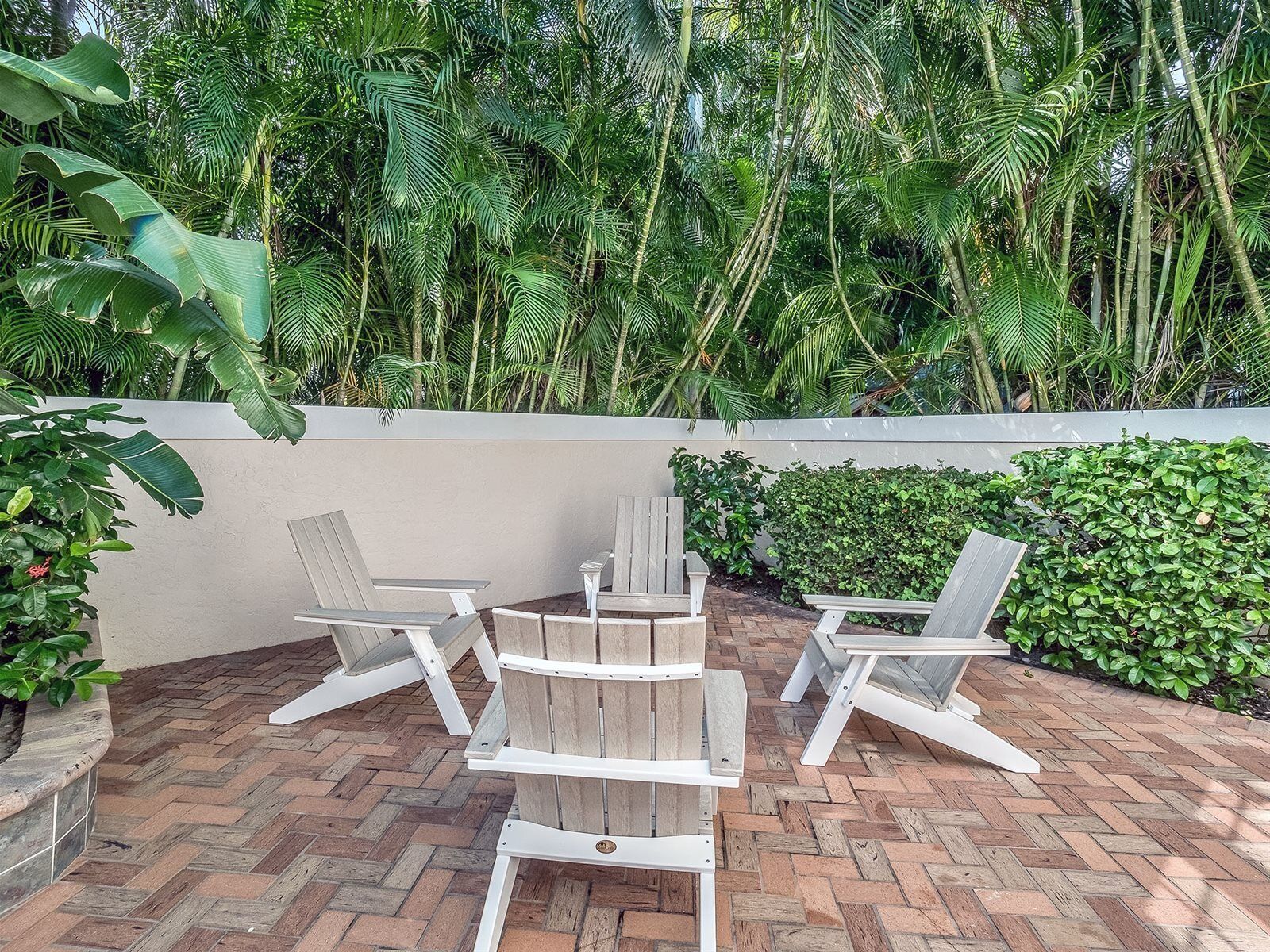 An outdoor patio with wooden chairs and a table, surrounded by lush greenery and a brick floor.