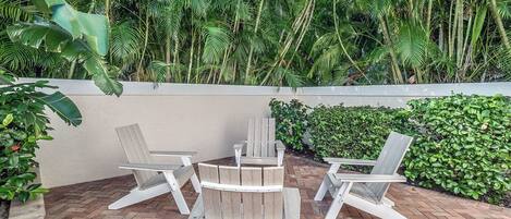 An outdoor patio with wooden chairs and a table, surrounded by lush greenery and a brick floor.