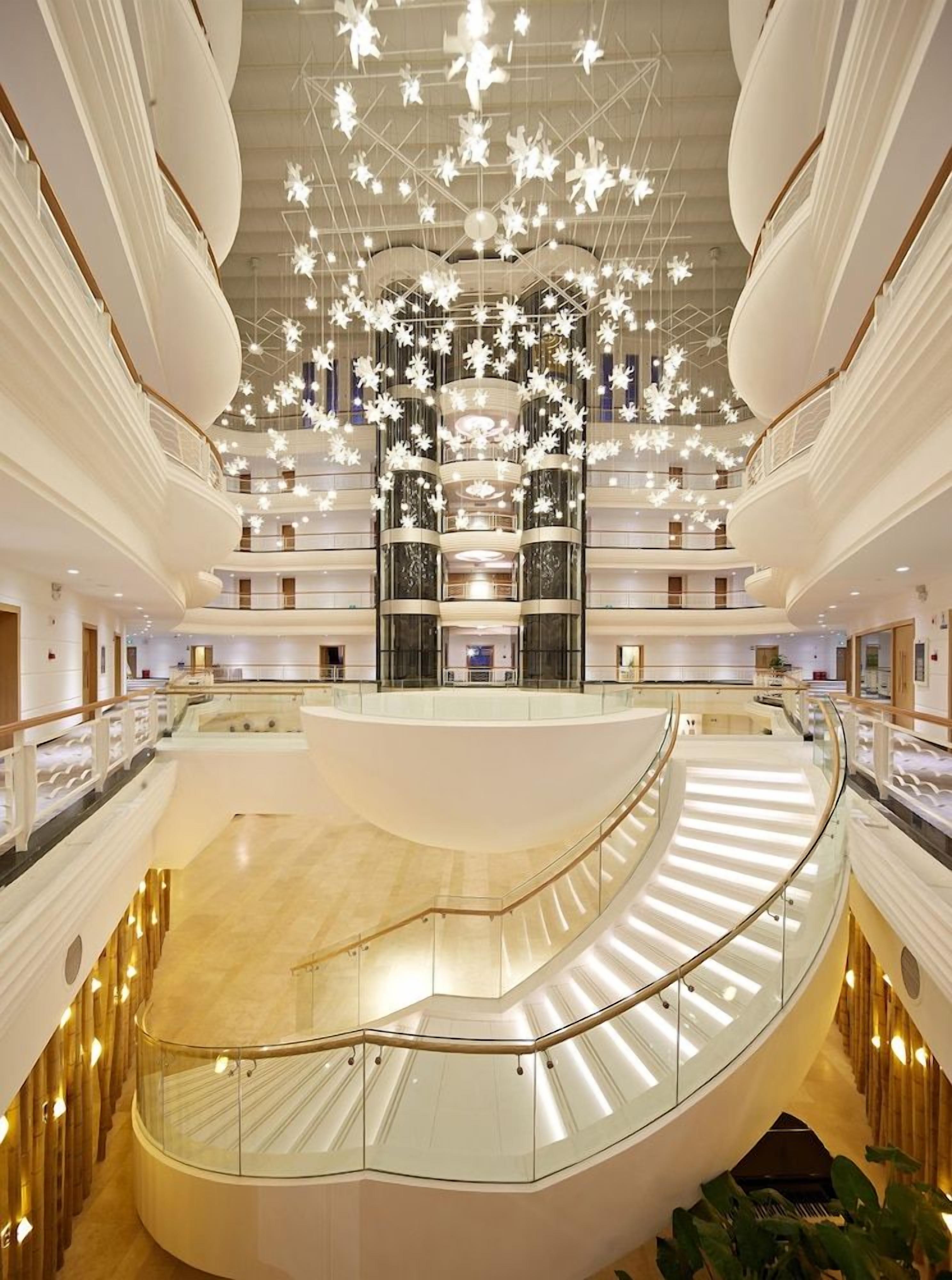 A multi-level atrium with a central staircase, glass railings, and a ceiling adorned with hanging light fixtures.