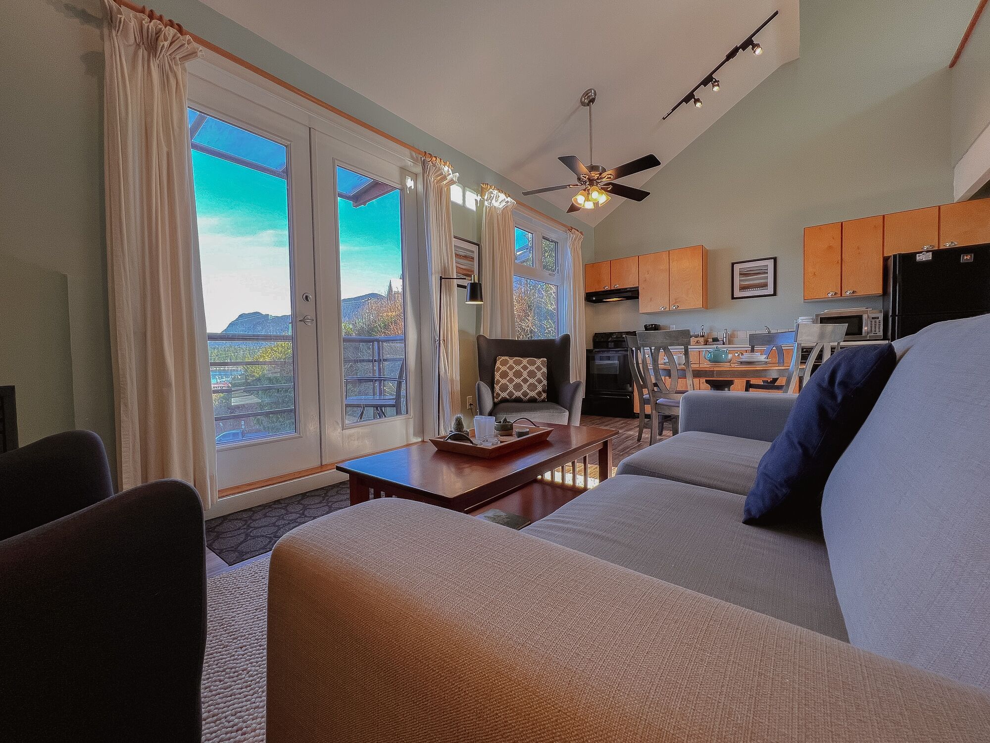 A living room with a sofa, a coffee table, and a dining area with a table and chairs. There is a kitchen area with cabinets and appliances.