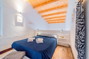 Une chambre avec un plafond en pente, un lit recouvert d’une couverture bleue, une table de chevet et une étagère fixée au mur.