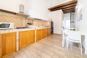 A kitchen with wooden cabinets, a microwave, stove, and a dining area with a table and chairs. - GuestHost - Tempio di Apollo - Ortigia Cozy Apartment (Syracuse)
