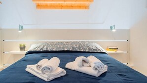 A modern bedroom with a wooden ceiling, a bed with dark blue bedding, two bedside tables with lamps, and a small potted plant on one table. - GuestHost - Tempio di Apollo - Ortigia Cozy Apartment (Syracuse)