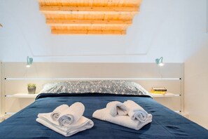 Une chambre moderne avec un plafond en bois, un lit recouvert d’une literie bleu foncé, deux tables de chevet avec des lampes et une petite plante en pot posée sur l’une des tables.