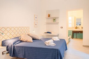 A bedroom with a bed, pillows, a bedside table, a shelf with books and a plant, and a bathroom visible through an open door. - GuestHost - Tempio di Apollo - Ortigia Cozy Apartment (Syracuse)