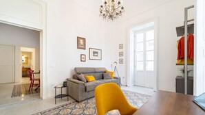 A living room with a sofa, a dining table, a chandelier, and a patterned rug. - Tempio di Apollo - Ortigia Historic Flat (Siracusa)