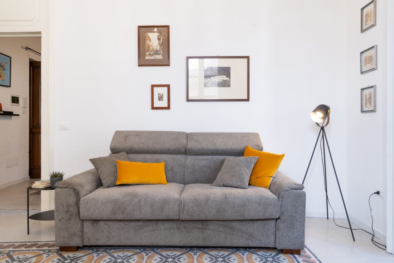 Un canapé gris avec deux coussins jaunes, un lampadaire et des tableaux encadrés accrochés au mur.