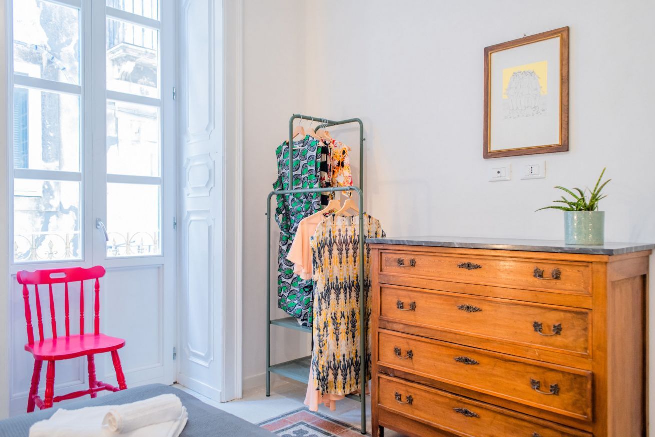 A room with a wooden dresser, a red chair, a clothing rack with various garments, a framed picture on the wall, and a potted plant.