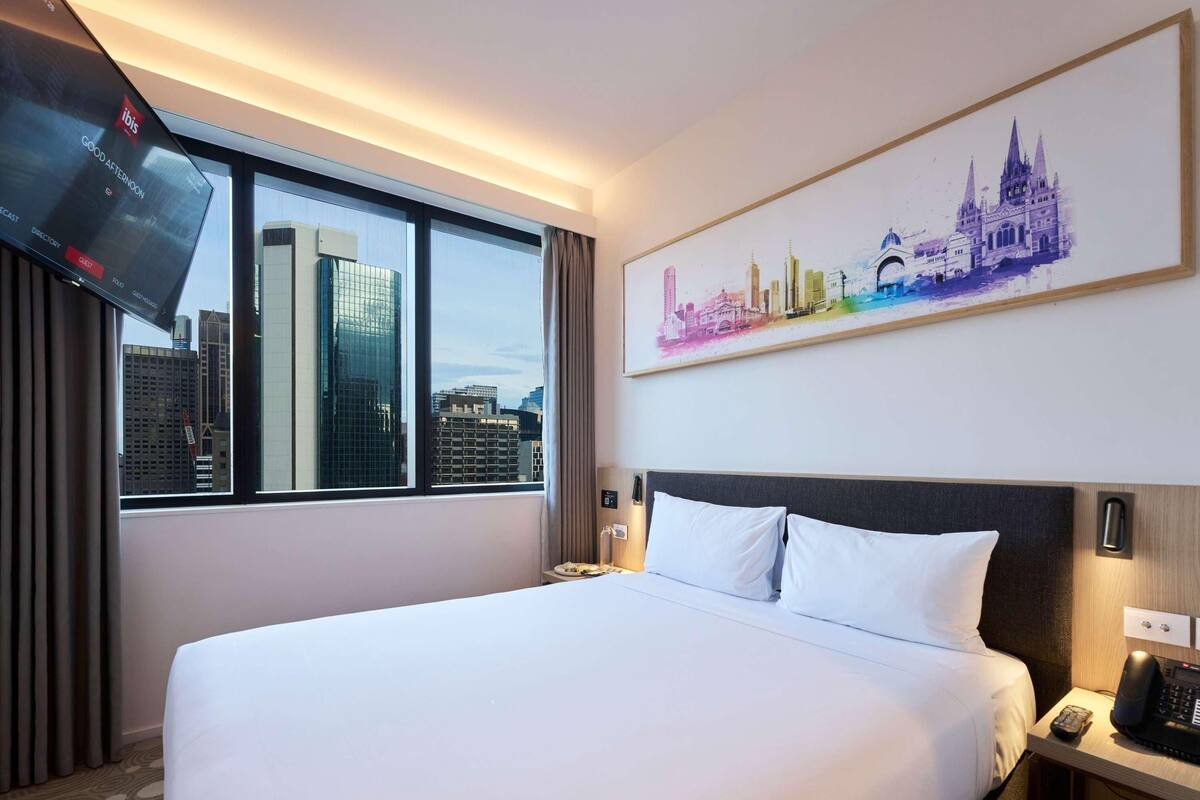 Une chambre d’hôtel avec un grand lit, un téléviseur fixé au mur, un tableau représentant un paysage urbain et une fenêtre donnant sur des immeubles imposants.