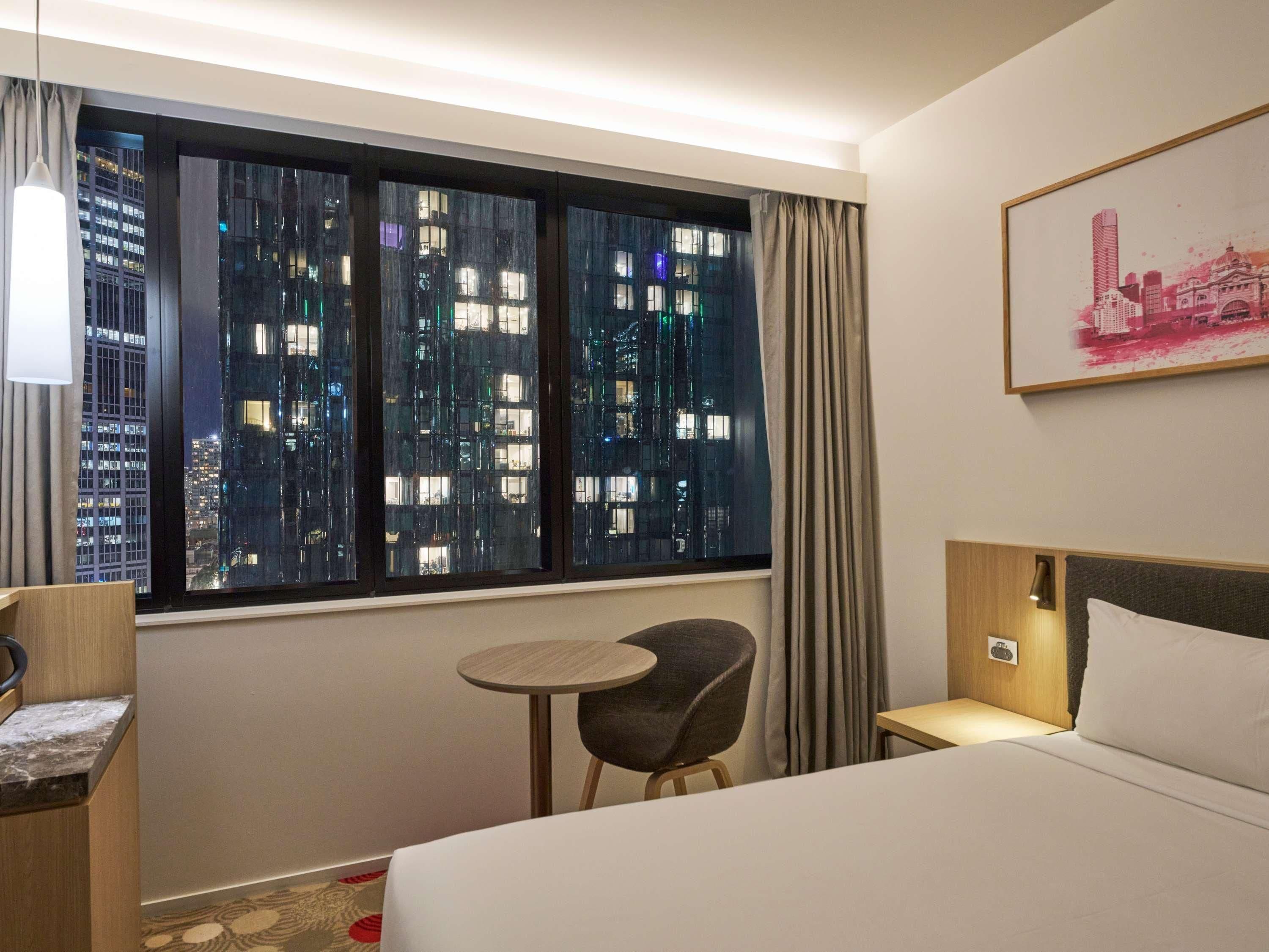 Une chambre d’hôtel avec une grande fenêtre, un lit, une chaise, une petite table et un tableau au mur.