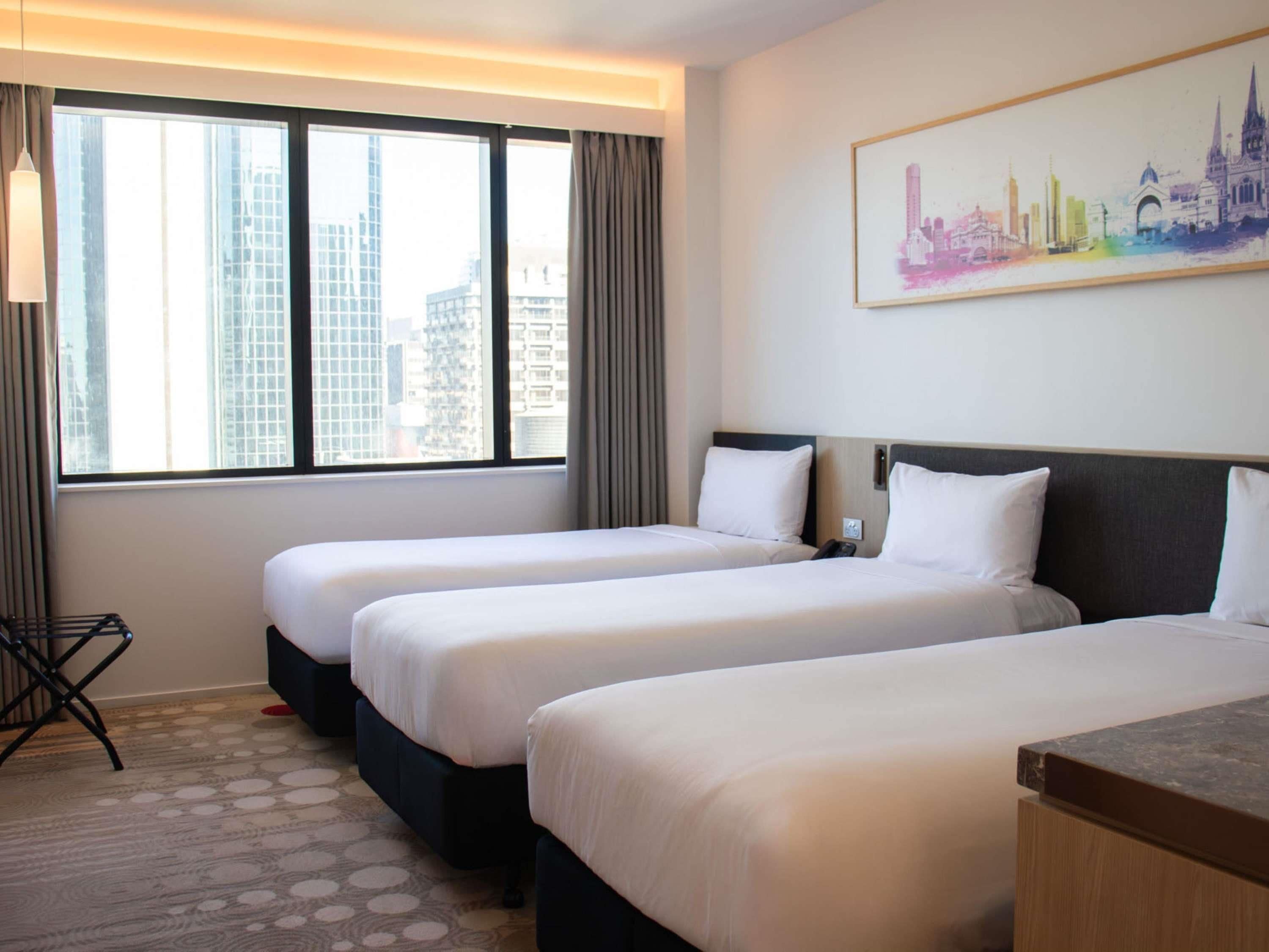 Habitación de hotel con dos camas, un cuadro de un paisaje urbano y vistas de edificios a través de la ventana.
