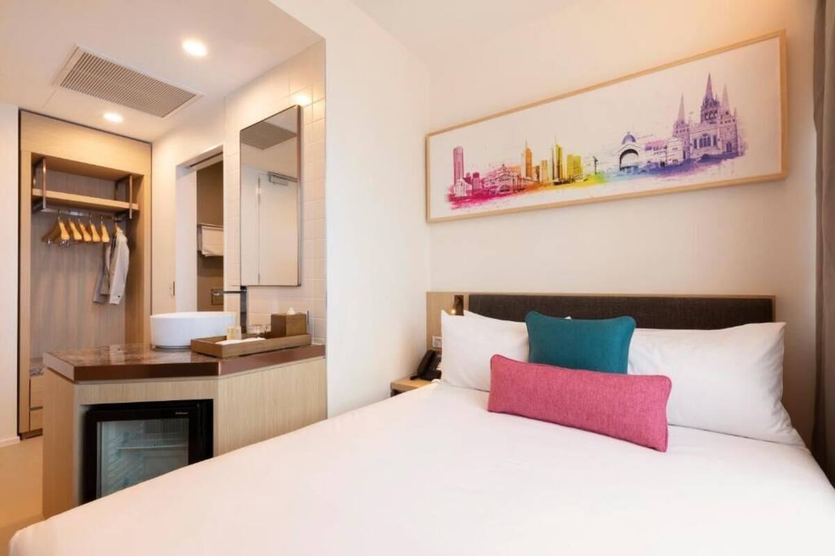 Une chambre d’hôtel comprenant un lit, une table de chevet, un miroir, une armoire et une œuvre d’art représentant un paysage urbain accrochée au mur.