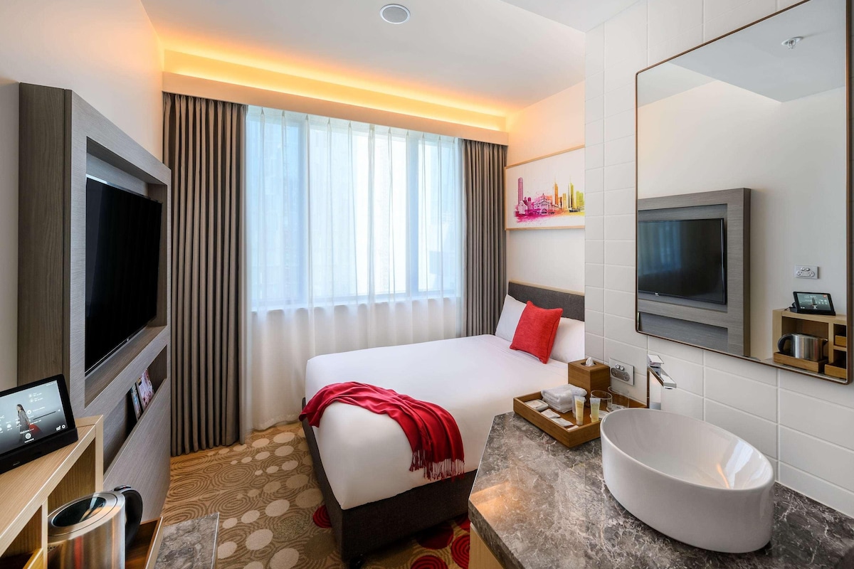 A hotel room with a bed, a TV, a large mirror, a sink, and a window with curtains.