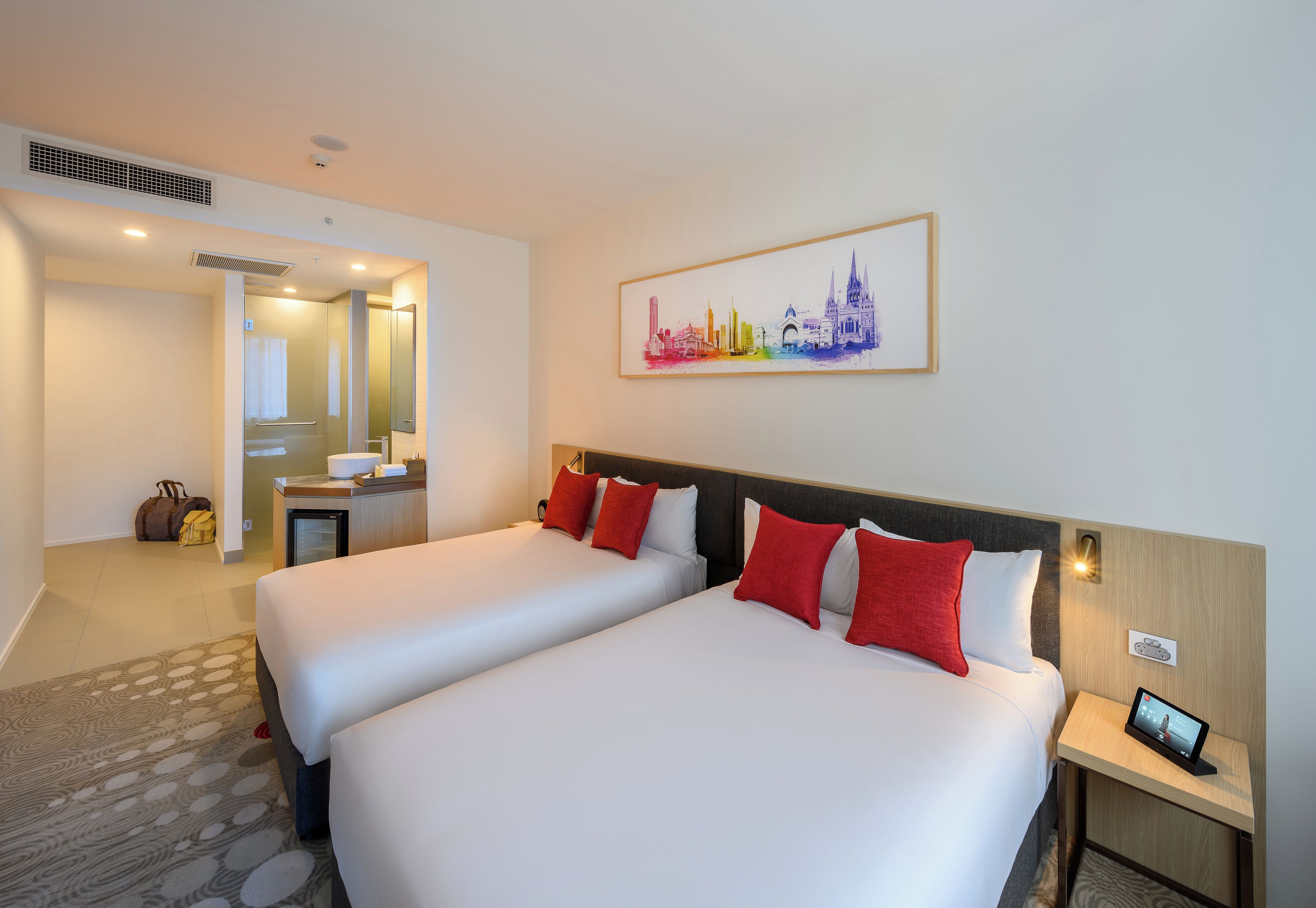 Habitación de hotel con dos camas, un escritorio pequeño, un espejo y una pintura de un paisaje urbano en la pared.