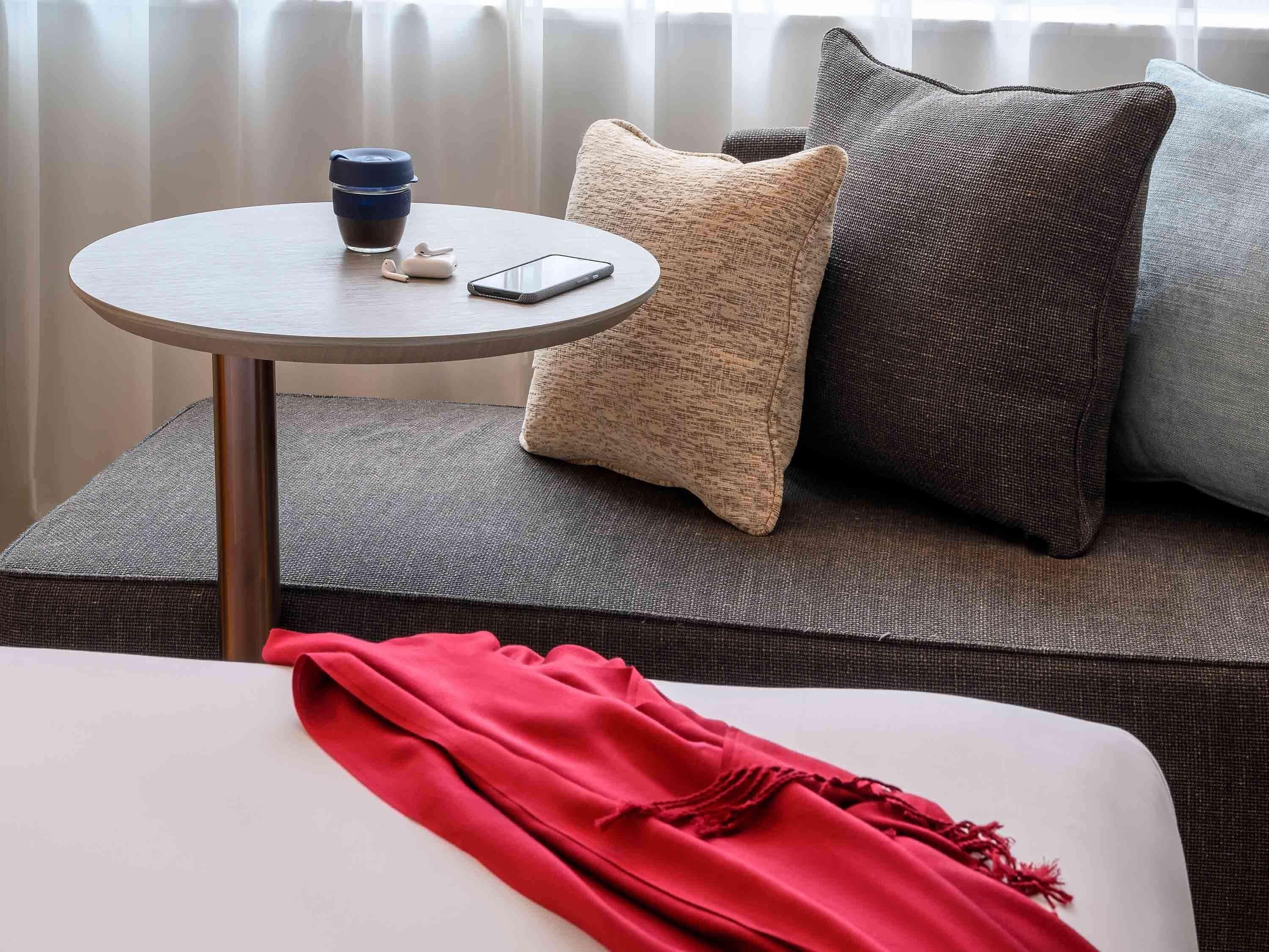 A modern hotel room with a round table, a smartphone, a coffee cup, and a red throw blanket on a sofa.
