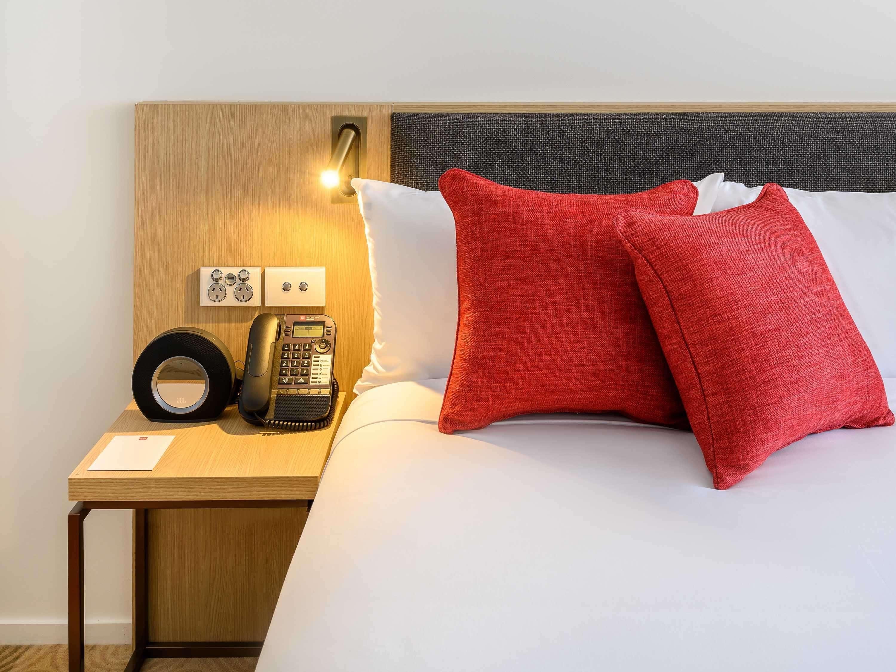 A hotel room with a wooden nightstand, a telephone, a power outlet, and a bed with red and white pillows.