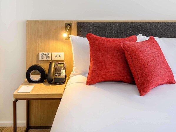A hotel room with a wooden nightstand, a telephone, a power outlet, and a bed with red and white pillows. - Ibis Melbourne Central (Melbourne)