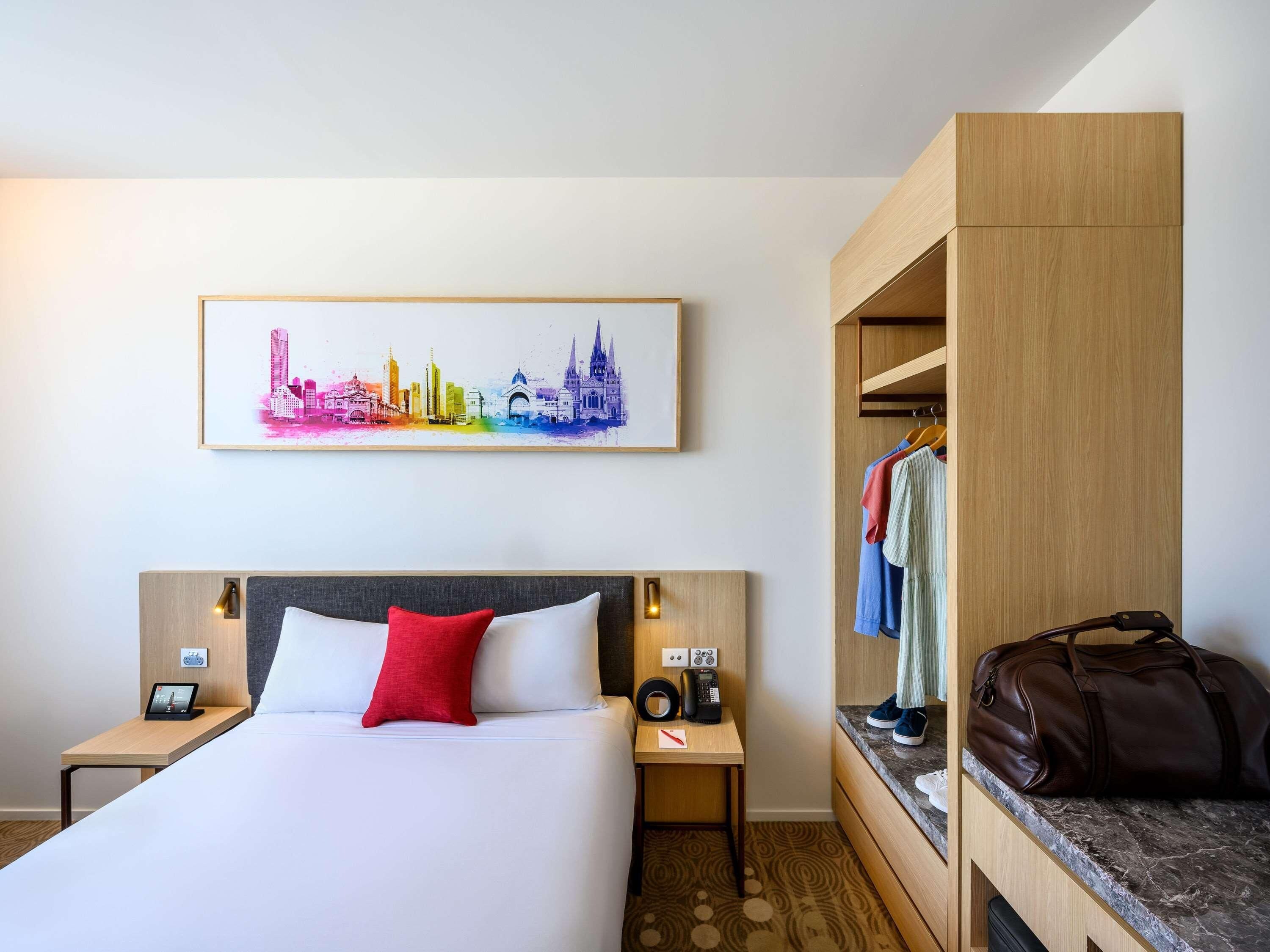 Une chambre d’hôtel comprenant un lit, des tables de chevet, une commode, une armoire, un porte-manteau et une œuvre d’art représentant un paysage urbain accrochée au mur.