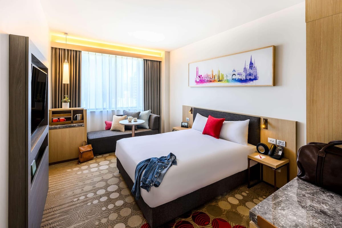Une chambre d’hôtel comprenant un lit, une table de chevet, une télévision, un coin salon et un tableau représentant un paysage urbain.