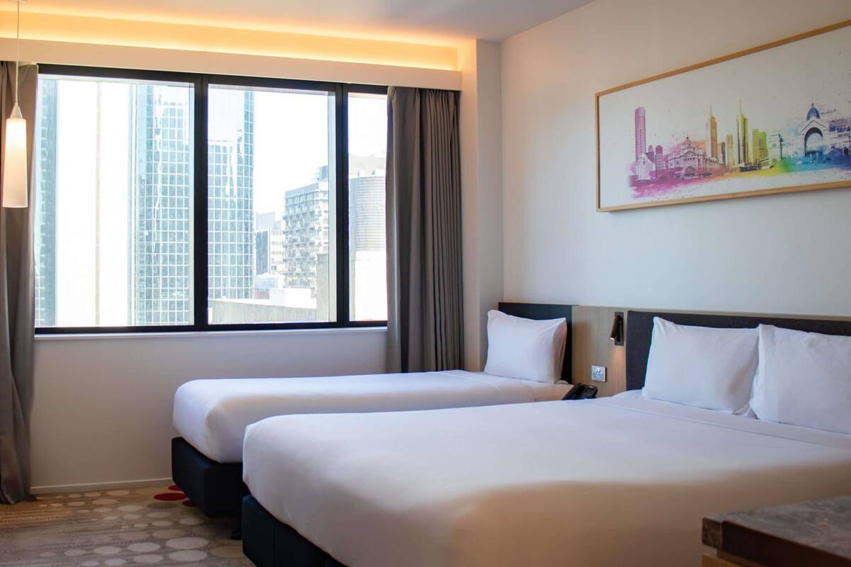 Une chambre d’hôtel avec deux lits, une vue sur la ville, un tableau au mur et un tapis à motifs.