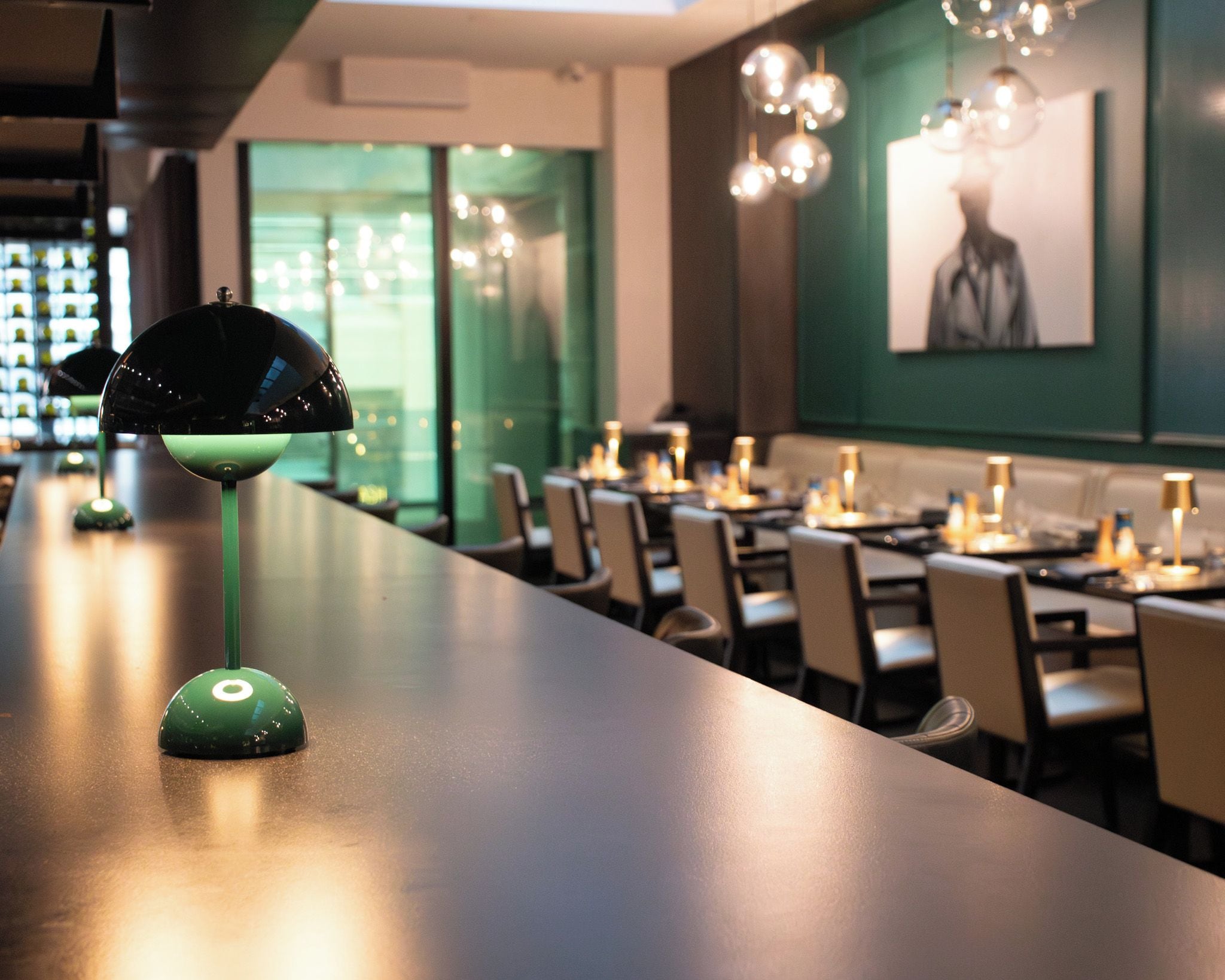 A bar area with a green lamp, tables, chairs, and a green wall with a framed picture.