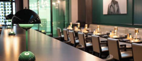 Zona de bar con una lámpara verde, mesas, sillas y una pared verde con un cuadro enmarcado.