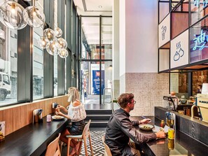 A modern restaurant interior with a long counter, hanging light fixtures, and a view of the street outside. - Ibis Melbourne Central (Melbourne)