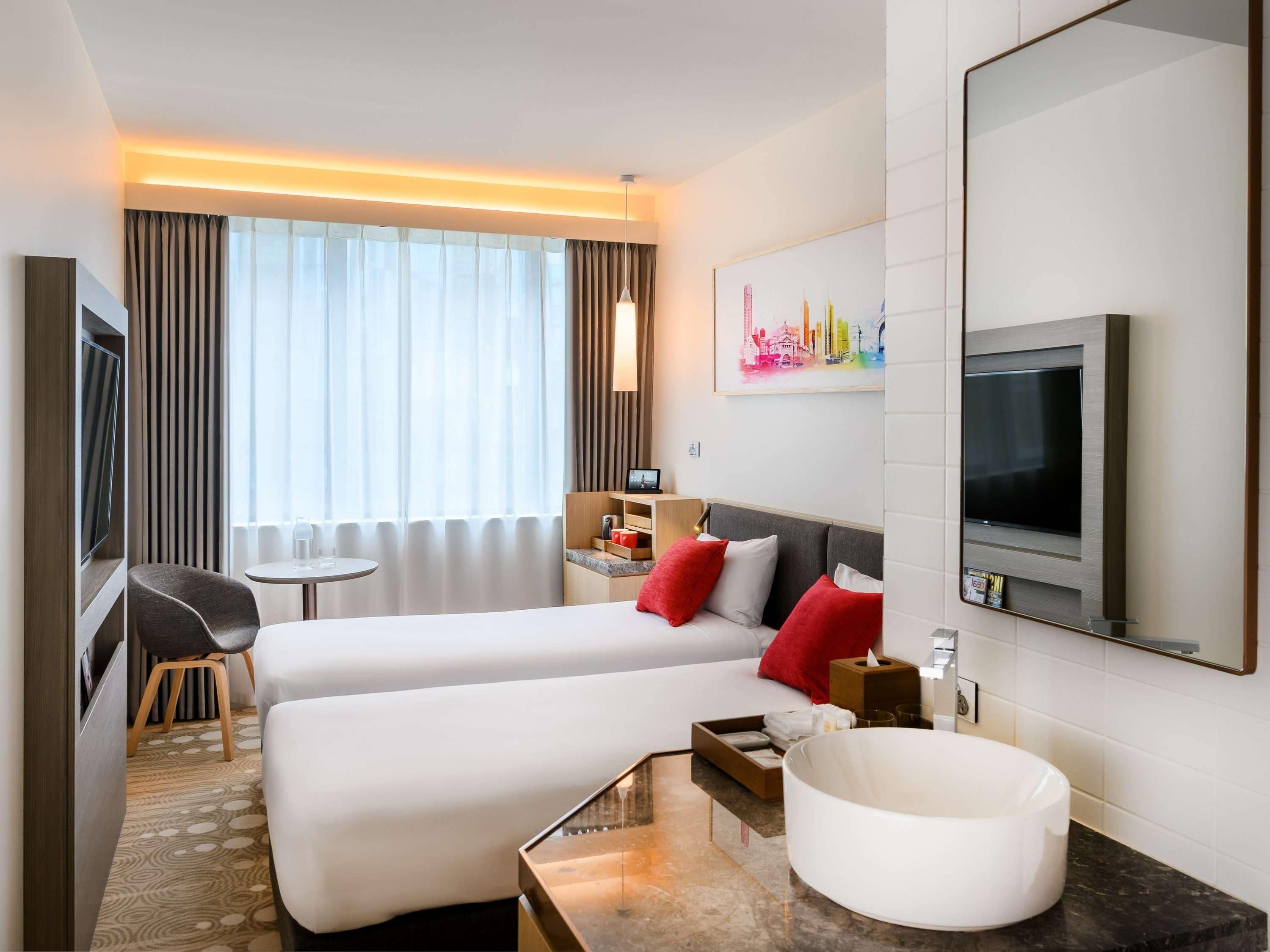 Habitación de hotel con dos camas, televisión, escritorio, silla, mesita de noche, espejo y un gran ventanal con cortinas.