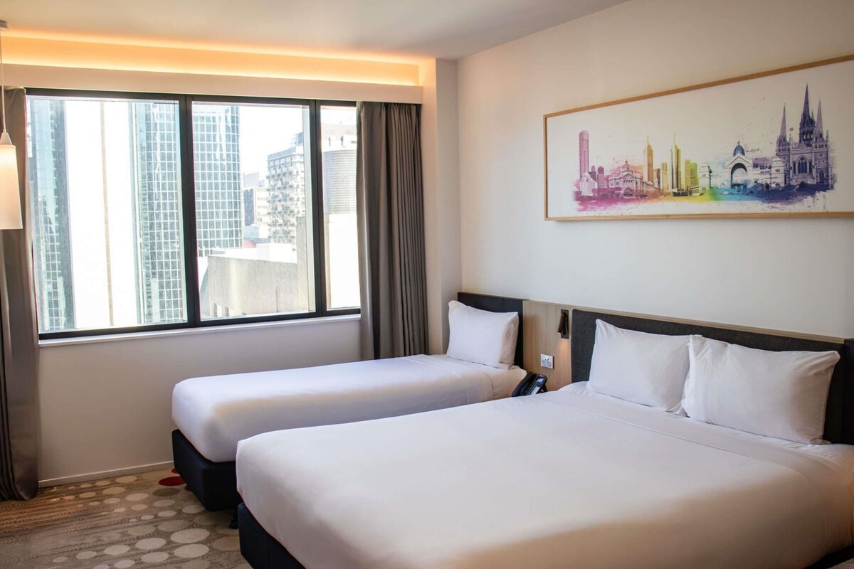 A hotel room with two beds, a cityscape view, and a painting on the wall.