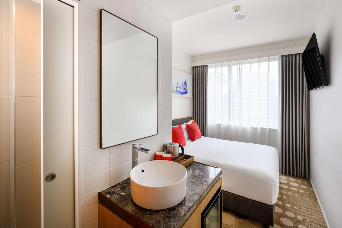 A hotel room with a bed, a large mirror, a round sink, a small table with a cup, and a window with curtains.