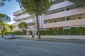 A multi-story residential building with balconies, a car parked in front, and a tree in the foreground. - New! Cozy APT x5 Near the Beach - Garage Included (Lignano Sabbiadoro)