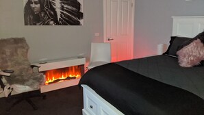 A bedroom with a bed, a chair, a white door, and a fireplace. - Country Luxury. Delux Queen Bed, Full Bathroom. Private Unit. Massage chair. (Scarsdale)