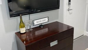 Ett rum med en platt-TV som är monterad på väggen, en byrå i trä med lådor, en flaska vin och ett glas på byrån.