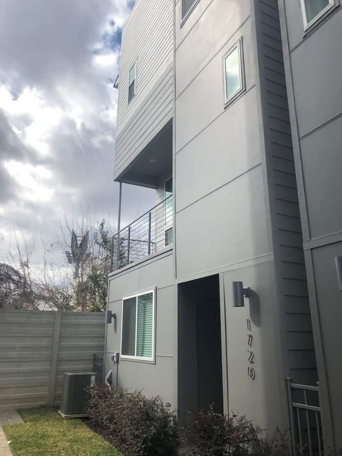 A modern multi-story building with a grey facade, a balcony on the second floor, and a small garden area in front.