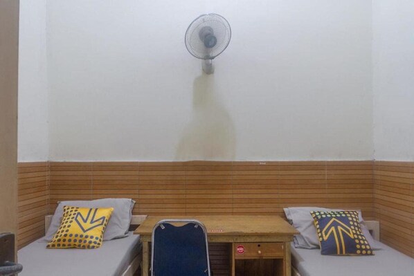 A room with two beds, each with a pillow featuring a geometric design. A small table is in the center, and a wall-mounted fan is above the beds.