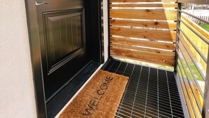 Eine schwarze Tür mit Fensterteil, eine Holztür mit Lamellen und eine Fußmatte auf einem Metallgitter.