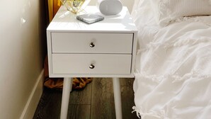 Ein Schlafzimmer mit einem weißen Bett, einem Nachttisch mit Lampe, einer kleinen Pflanze und einem weißen Nachtschränkchen mit Schubladen.