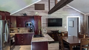A modern kitchen with dark wood cabinets, a stainless steel refrigerator, a microwave, and a large island with a sink. There is a dining area with a wooden table and chairs, and a flat-screen TV mounted on the wall. - Chalet lacbrome (Knowlton)