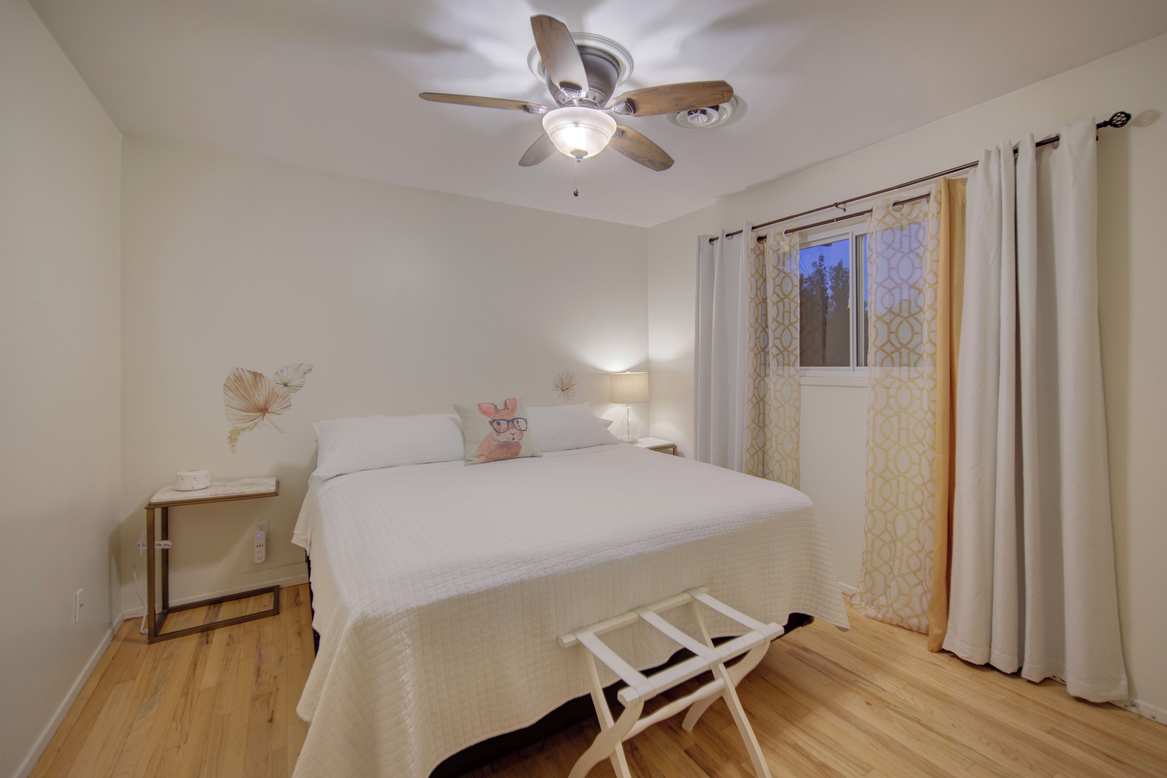 A bedroom with a bed, a nightstand, a bedside table, a ceiling fan, and a window with curtains.