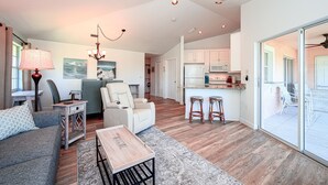 Amplia sala de estar con sofá, mesa de centro, sillón y lámpara. Zona de cocina con refrigerador, microondas y mostrador tipo bar. La habitación cuenta con un amplio ventanal corredero que da a un patio exterior.