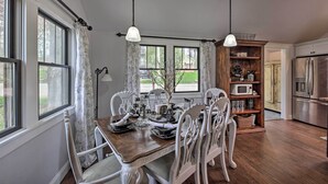 Uma sala de jantar com uma mesa de madeira e cadeiras brancas, um lustre e vista para o exterior através de grandes janelas.